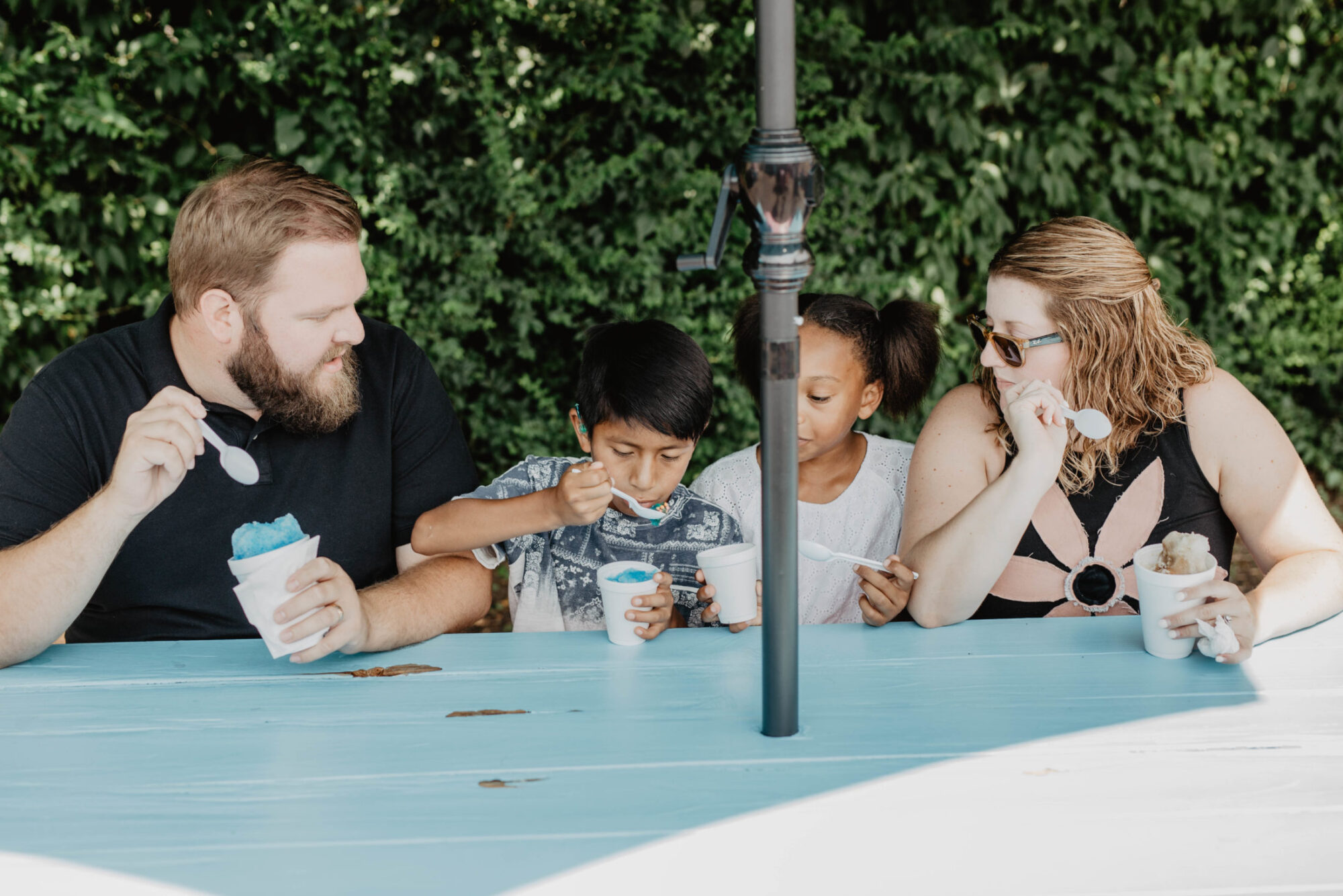 Two adults and two foster children sharing a happy moment at a picnic table with snow cones, in front of green foliage.