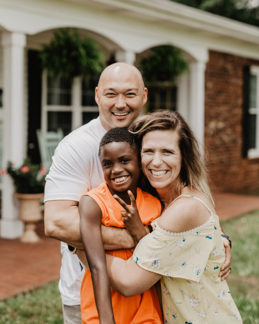 A happy multiracial foster family of three, including a smiling adult couple embracing a young boy who is making a peace sign, in front of a home.