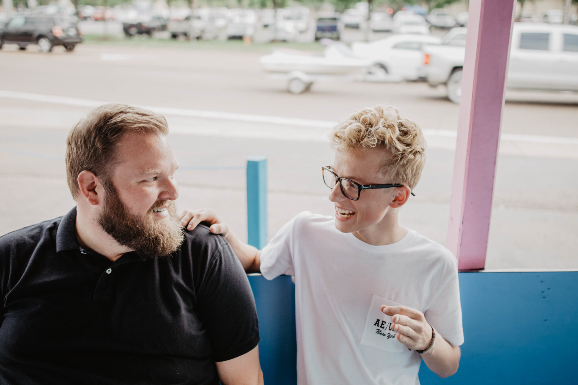 Two smiling people, one adult and one teenager with glasses, with teenager putting hand on adult's shoulder.
