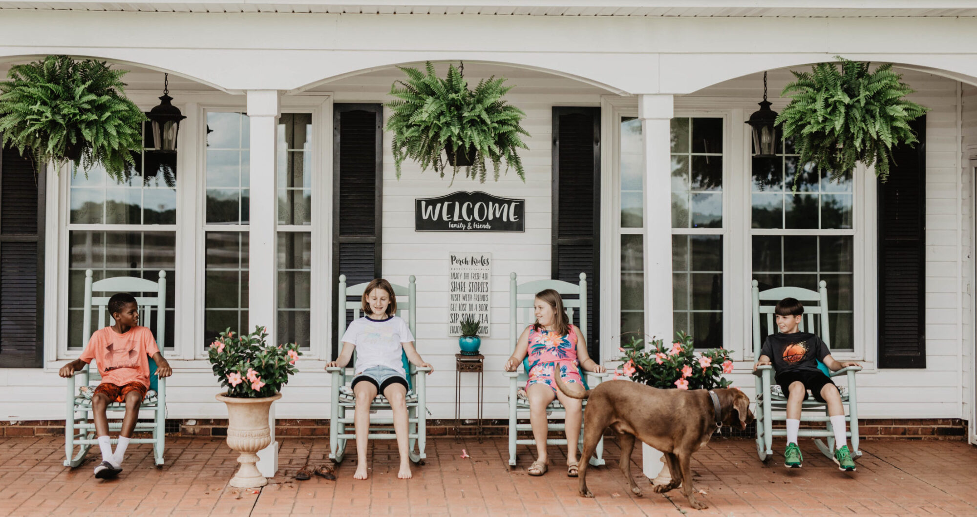 Four children sitting on white rocking chairs on the front porch of a group home.