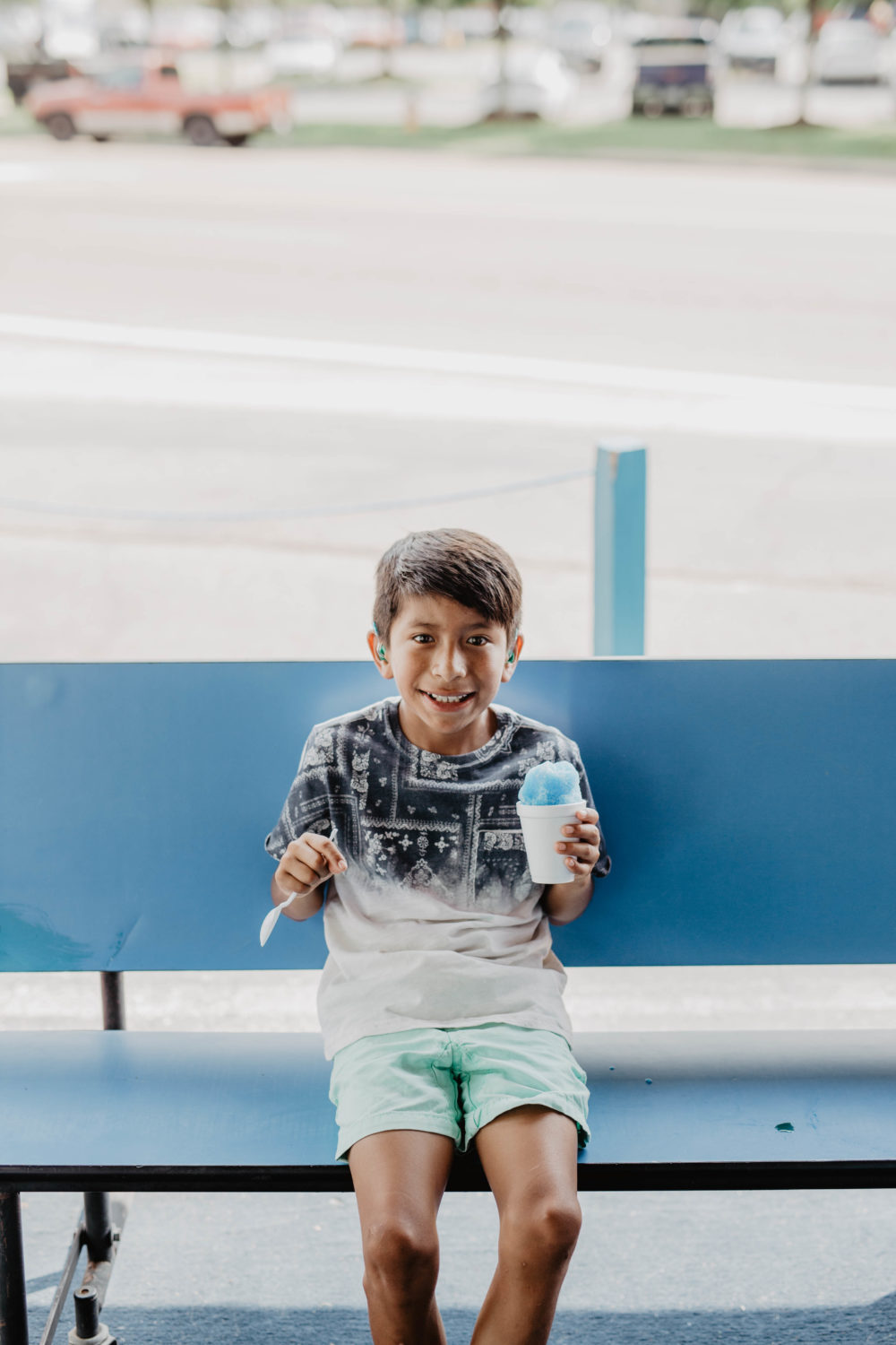 A smiling child on a blue bench with a spoon in one hand and a snow cone in the other.