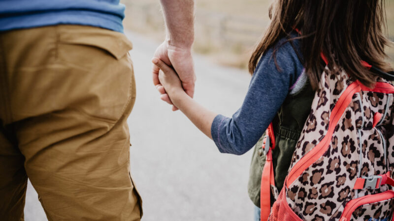 A dad and his daughter with a backpack holding hands.