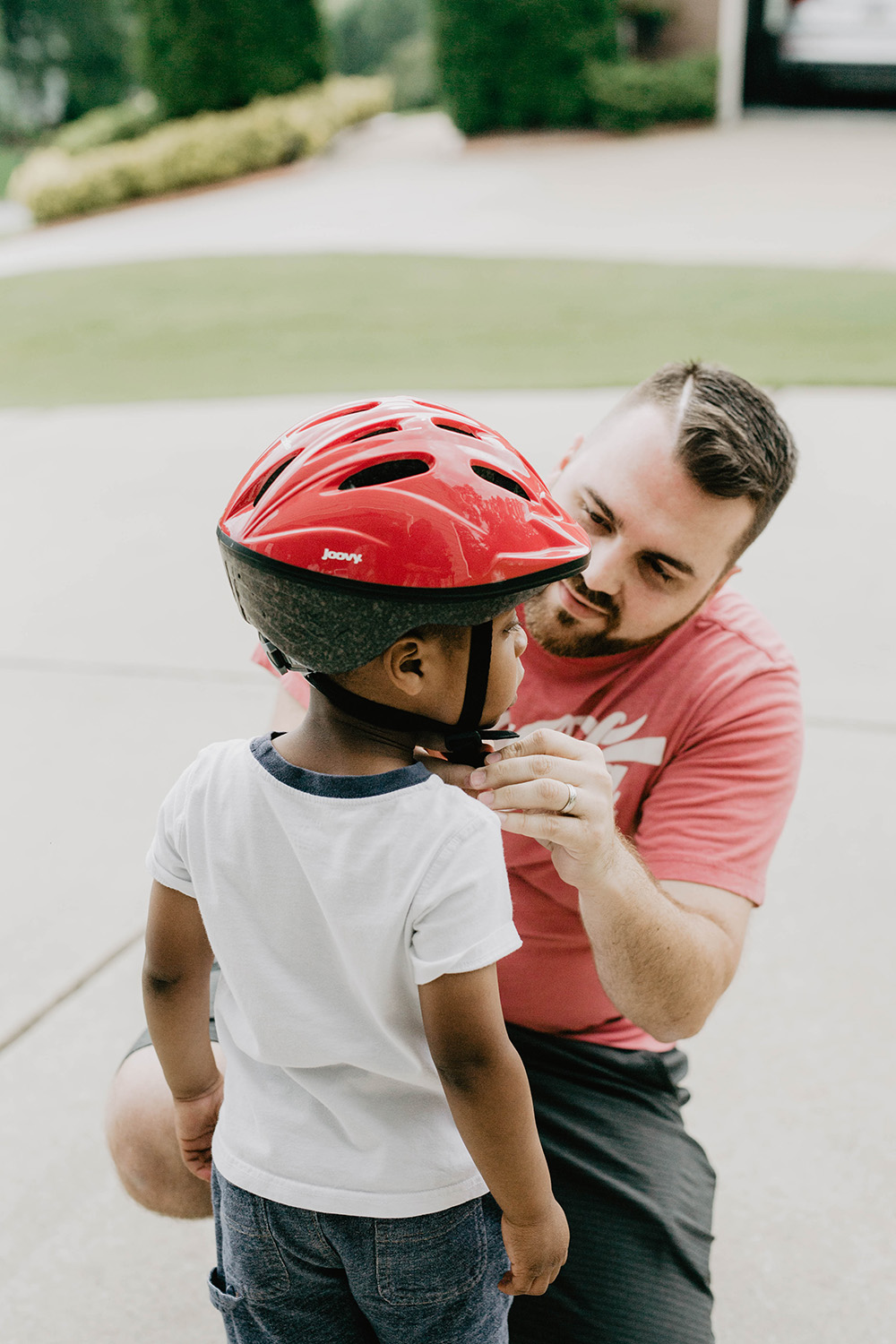 An adult male kneeling and helping a young child with a red helmet, securing the strap, in a driveway.