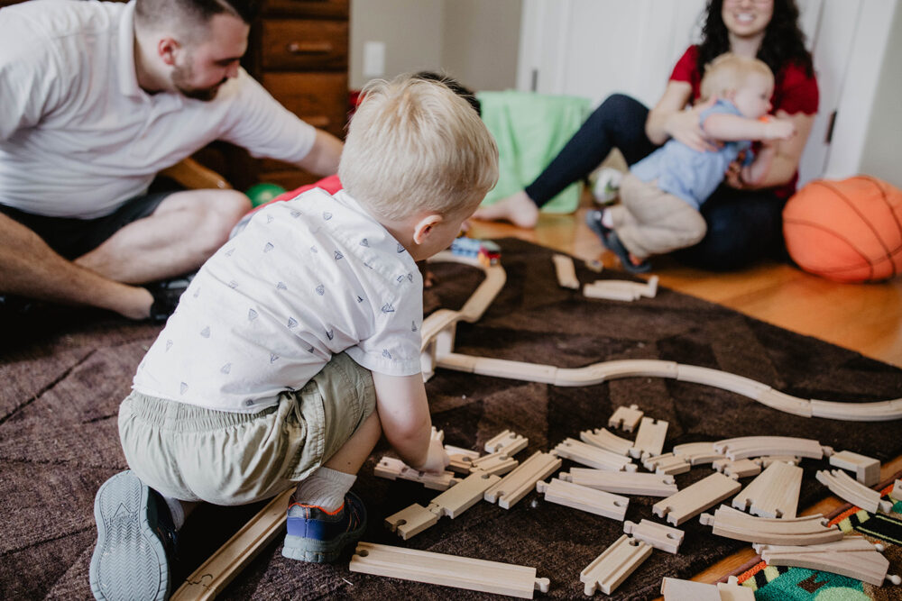 A young blonde foster boy playing on the floor with a wooden train set, while adults and another child are visible in the background.
