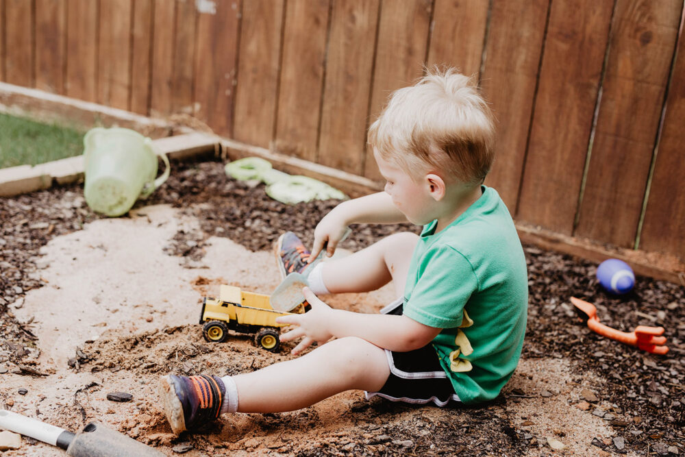 A young blonde boy in a green shirt playing happily in the dirt with a toy dump truck and shovel in a backyard.
