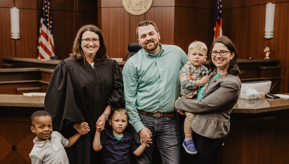 A happy family portrait in a courtroom with a couple, their three adopted children, and a smiling judge, finalizing the adoption.