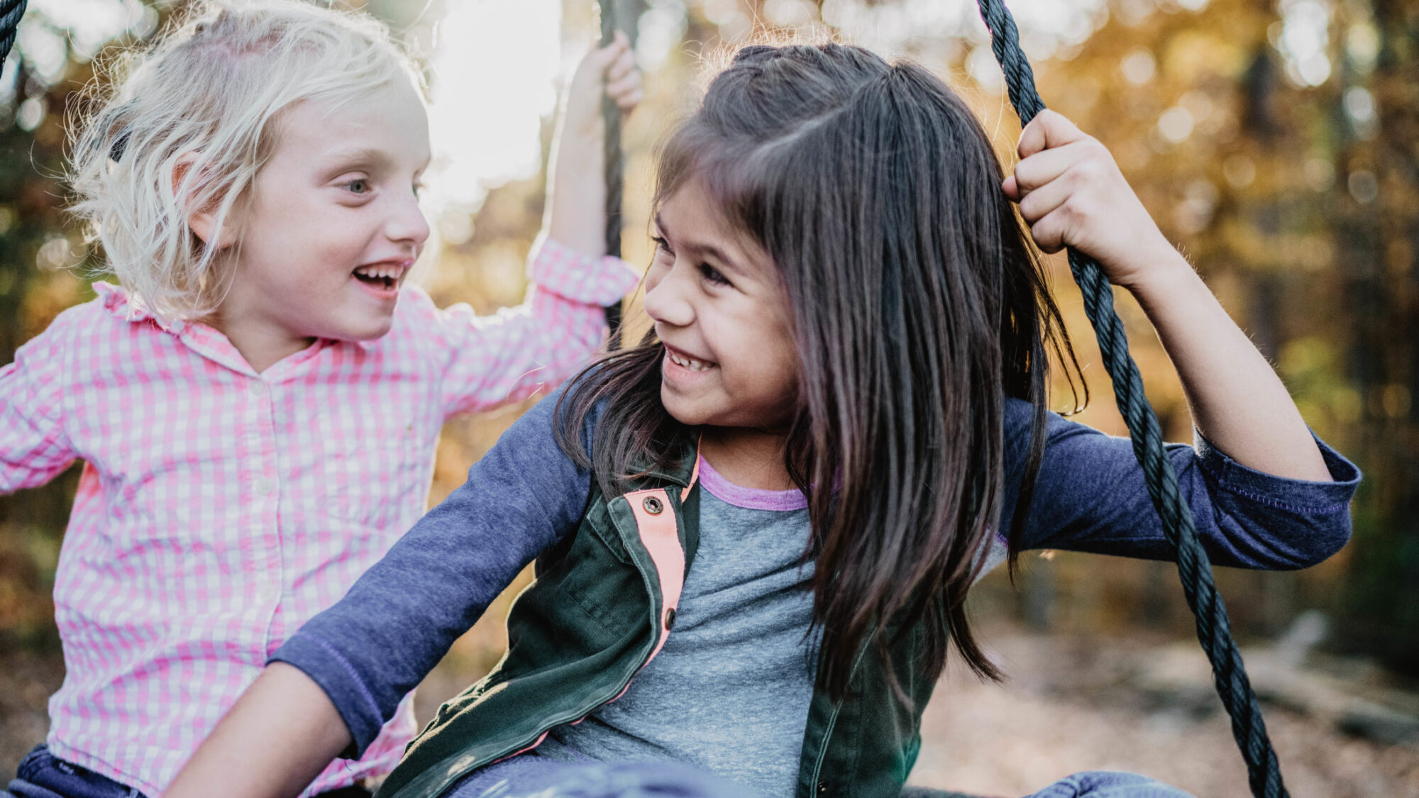 Two girls smiling at each other while swinging outside.
