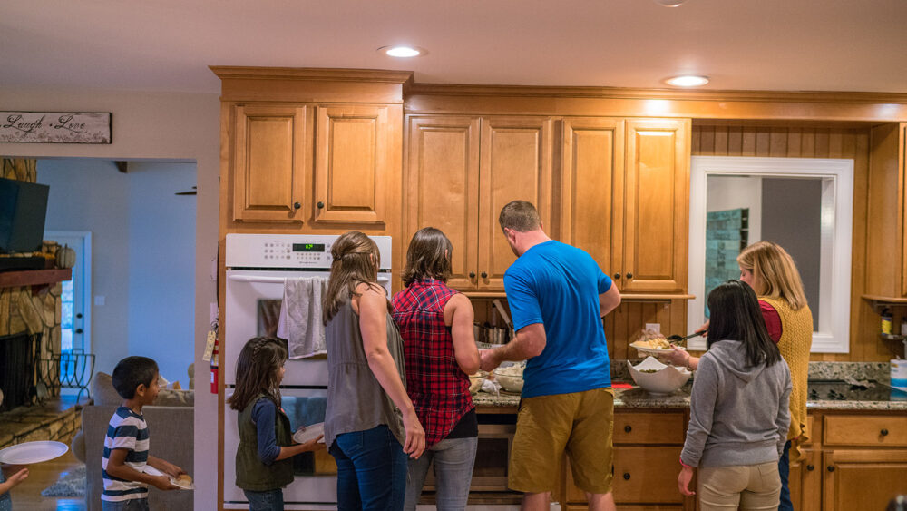 Multiple adults and young children in a modern kitchen, standing around countertops with food.