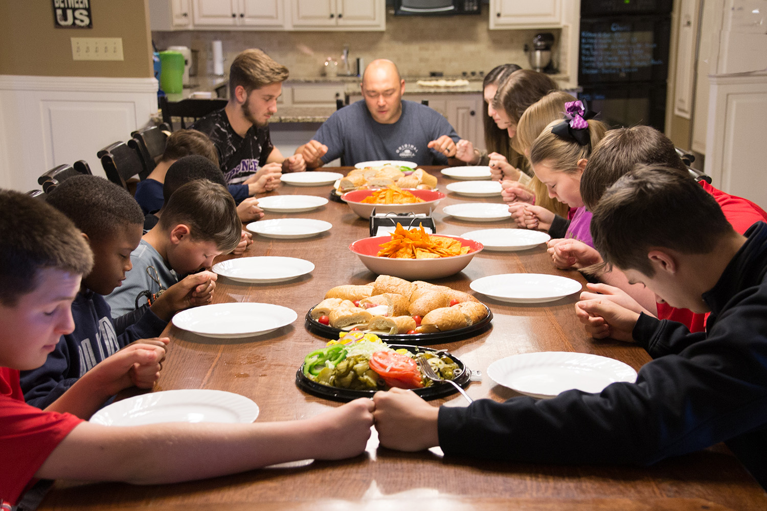 A large foster family praying at a long wooden dining table with food on it.