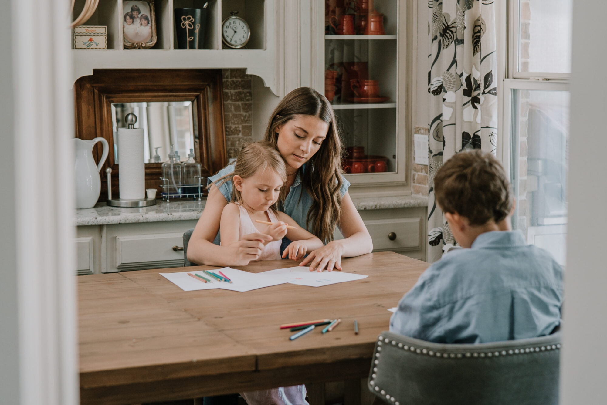 A foster mom helping a girl draw at a wooden table with boy sitting across as one of many quarantine activities.