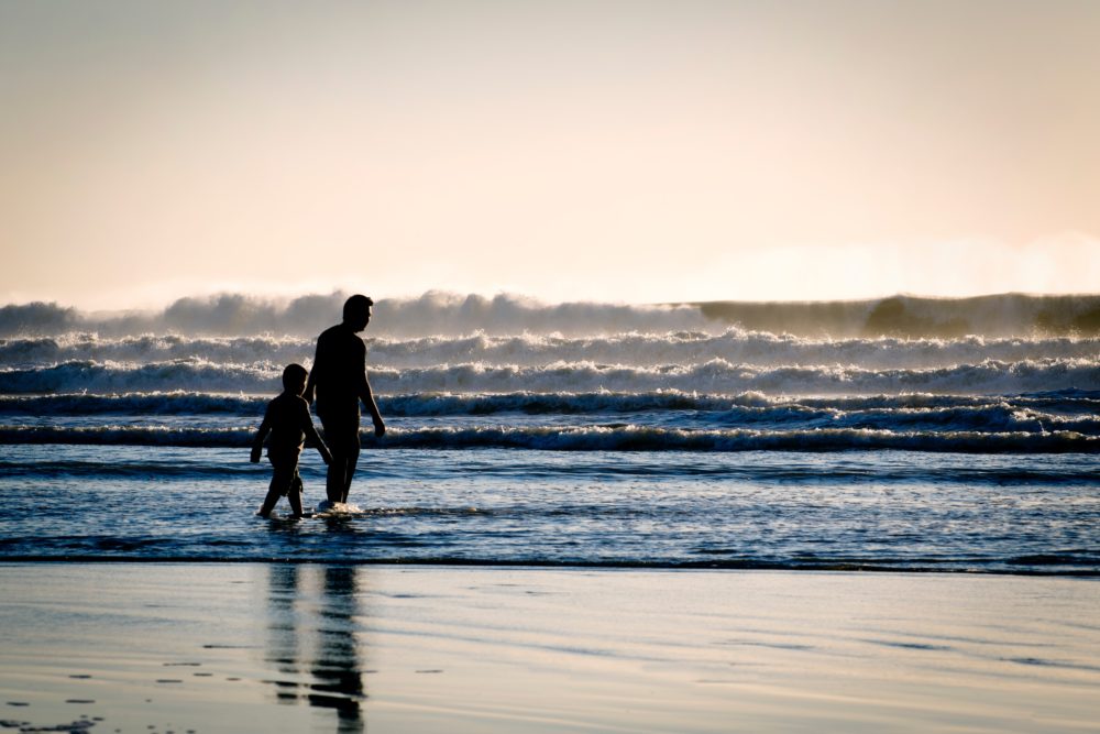 A father an son walking in shallow water on a beach.