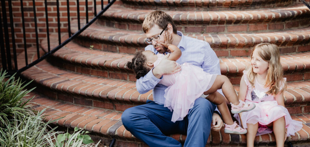 A father kissing a foster girl on brick stairs while another girl laughs.