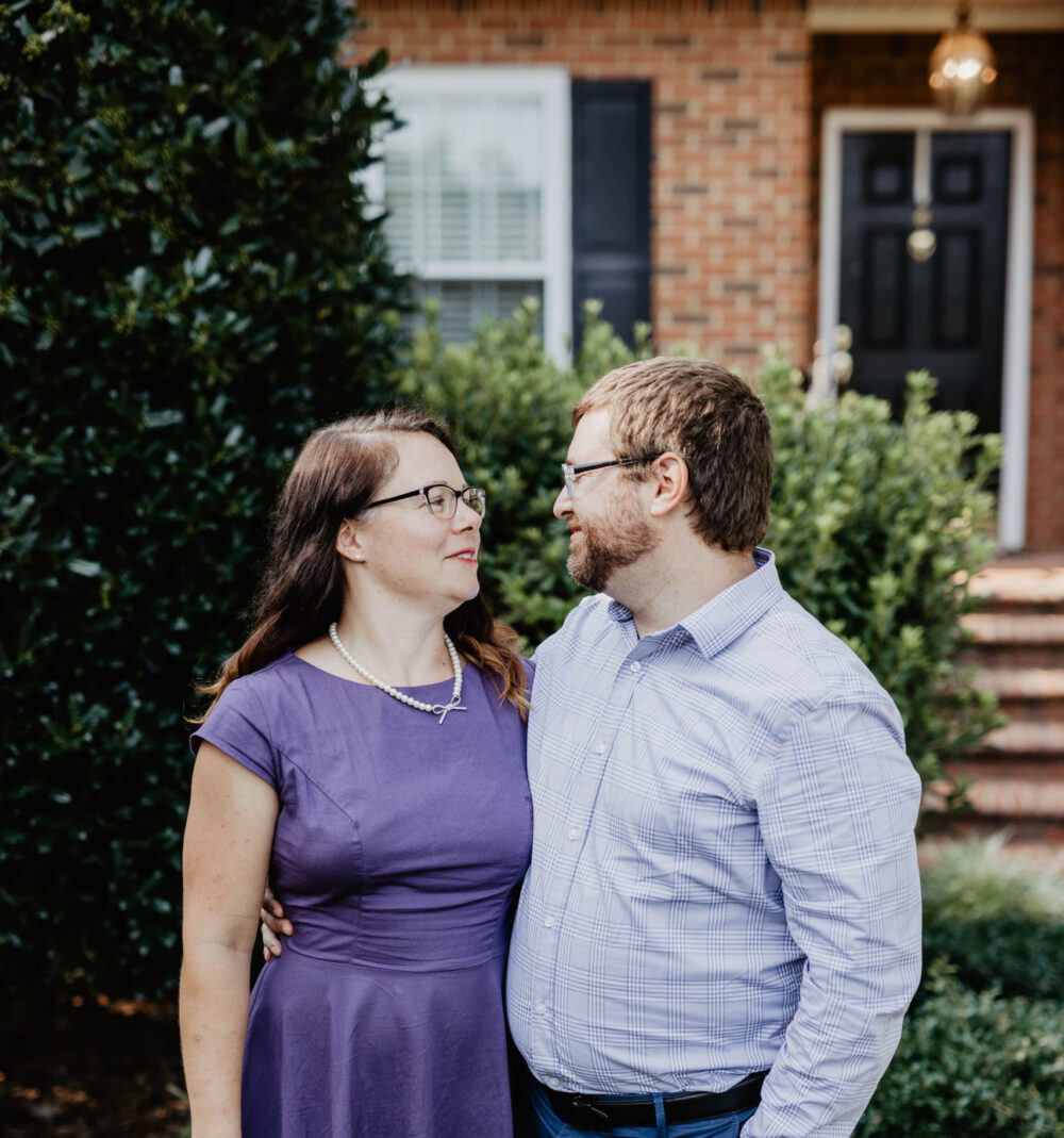 A man and woman looking at each other smiling in front of brick house and bushes.