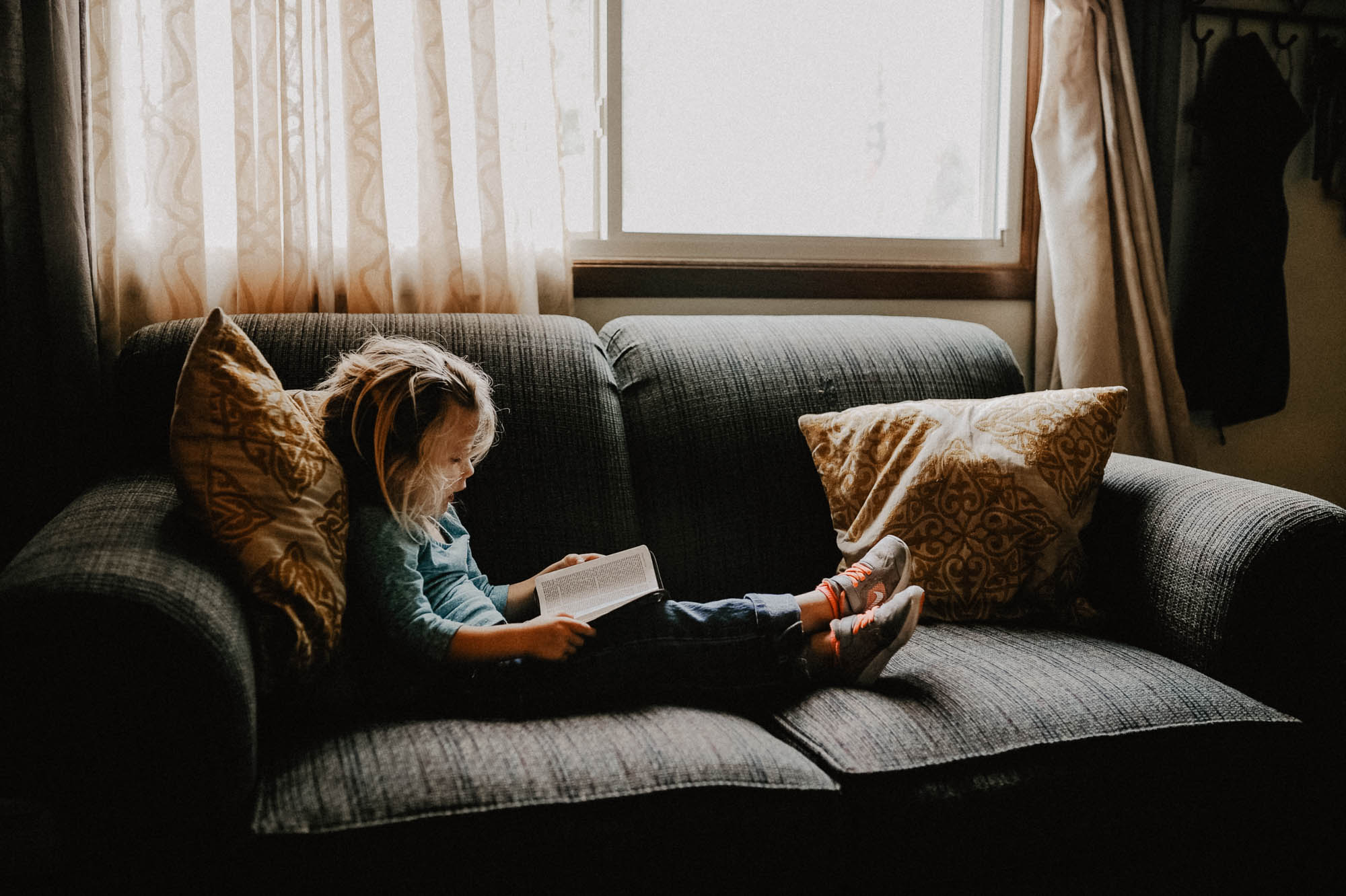 A girl sitting on a dark couch reading a book next to a bright window.