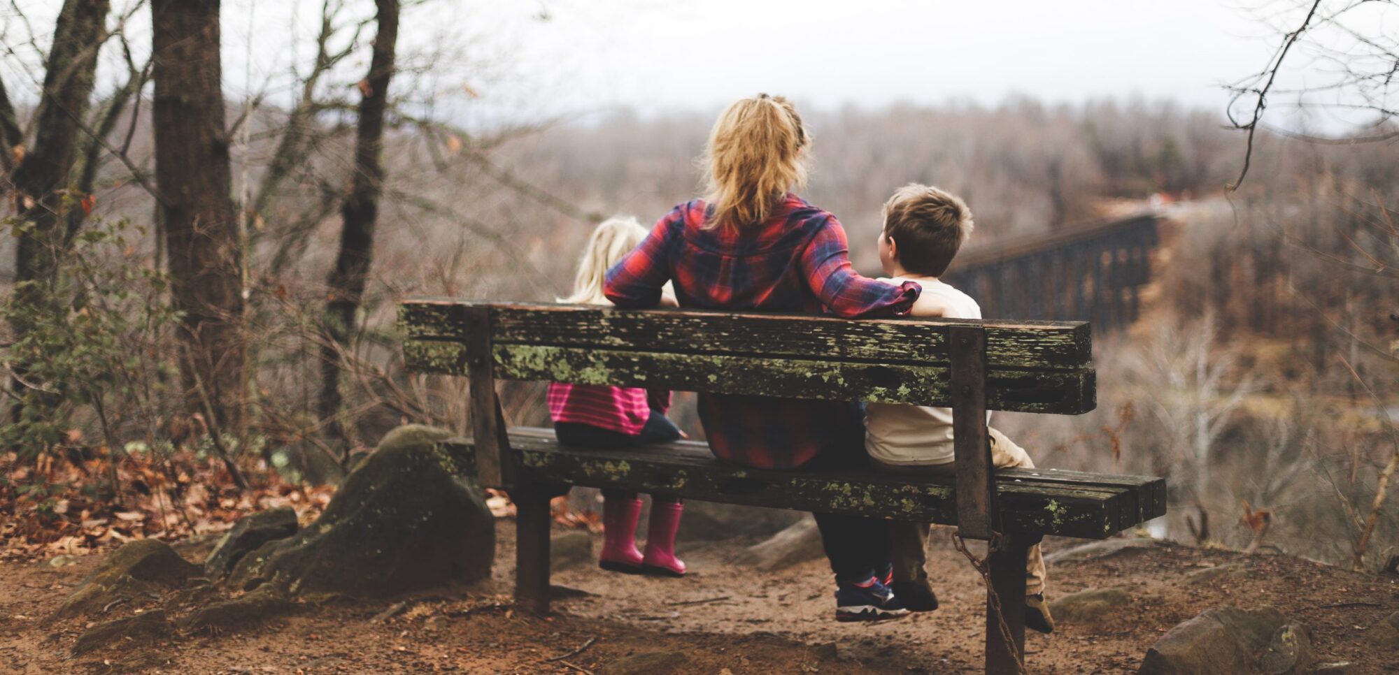 Foster mom practicing self-care while sitting on bench with two foster children in woods.