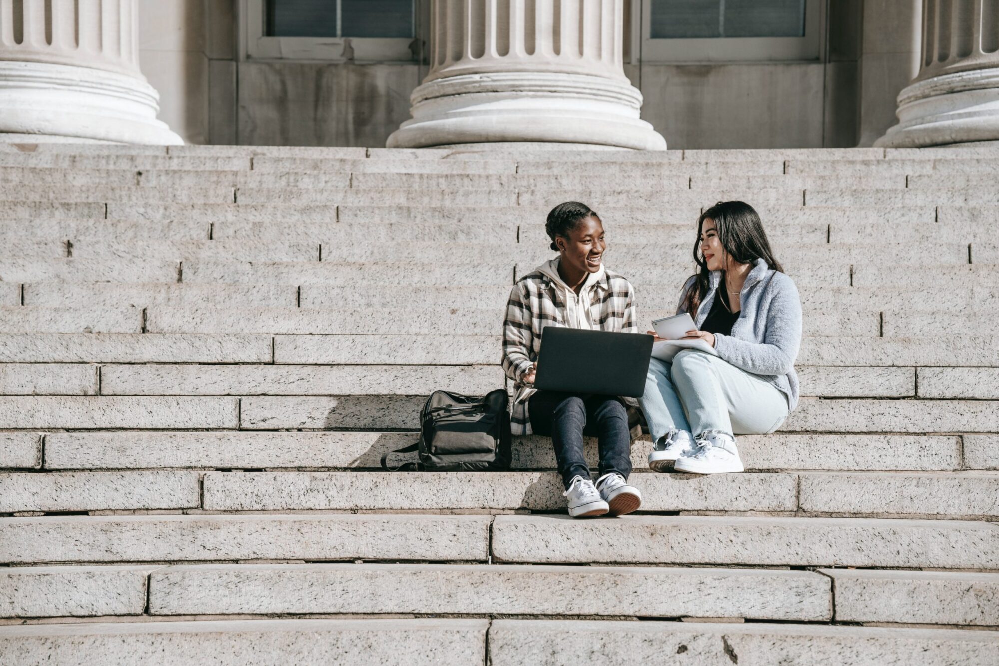 Two students sitting on stairs with a laptop and notebook.
