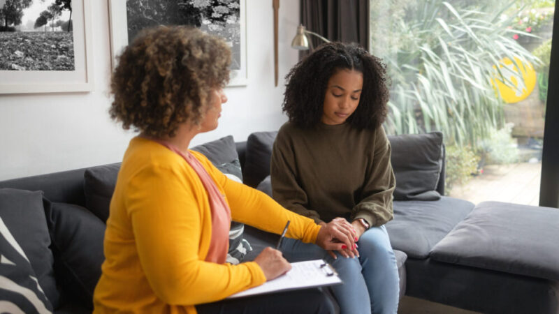 A woman putting her hand on a girl's hand while sitting on a couch together.