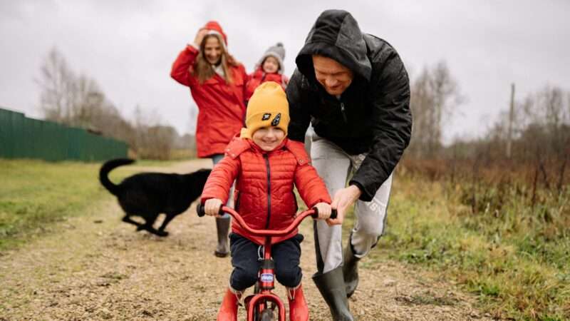A happy family outside on a dirt road with dad helping son ride a bike.