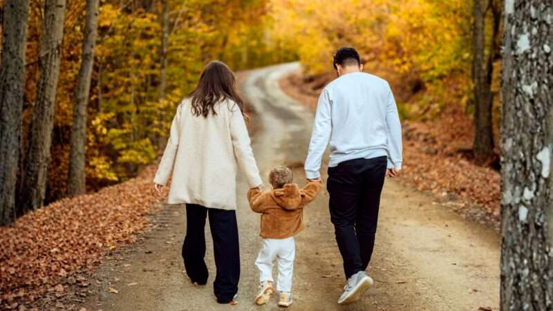 A dad and mom holding a child's hand on a dirt road in the woods during fall